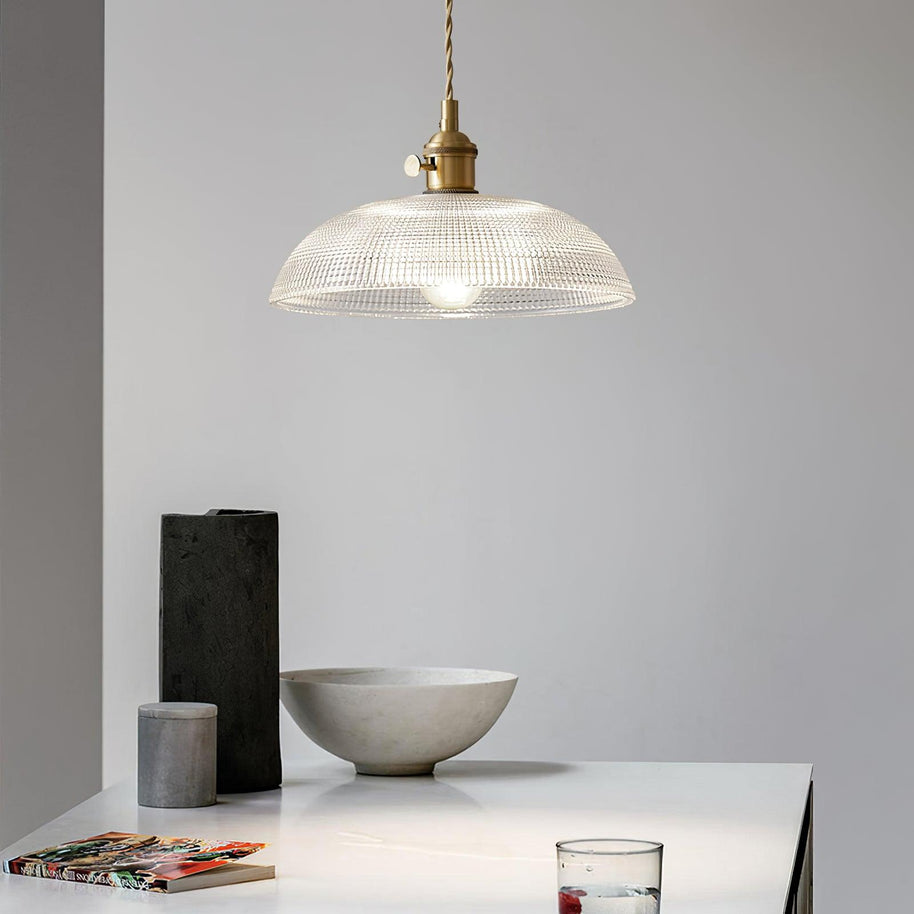Pendant light with a ribbed glass dome shade and a gold-toned lamp holder hanging above a table set with a bowl and dark ornaments.