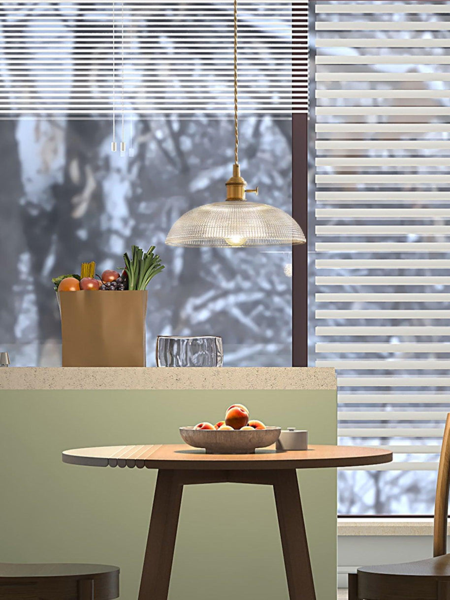 Pendant light with a ribbed glass dome shade and a gold-toned lamp holder hanging over a kitchen counter, seen through a window.