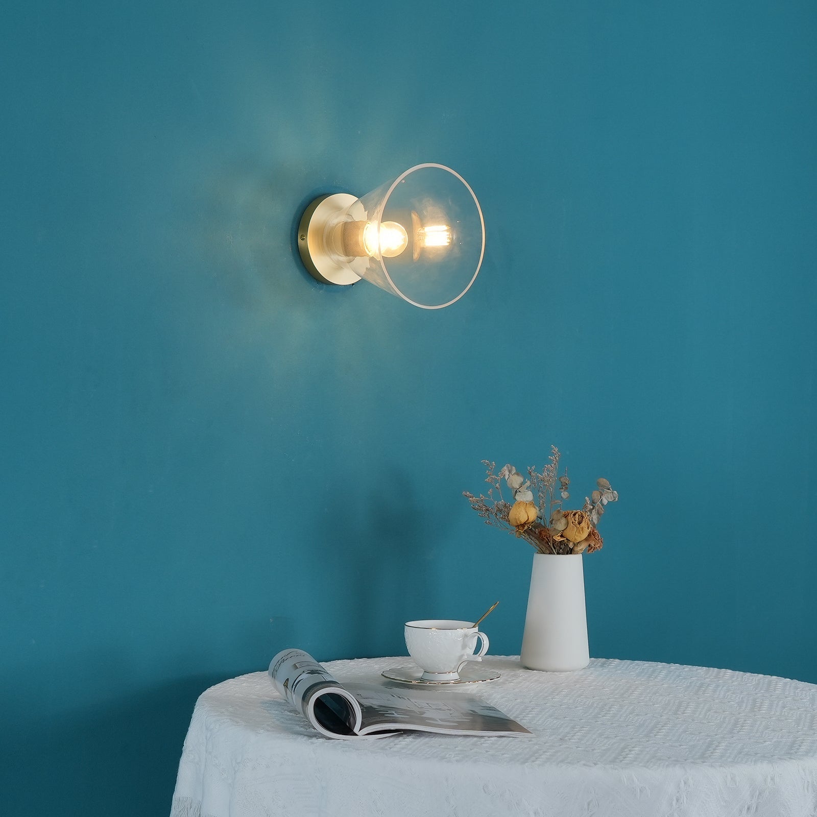 Light fitting with a round brass-toned back plate and a rounded glass shade, lit, against a teal background, with a table, cup and dried flowers below.