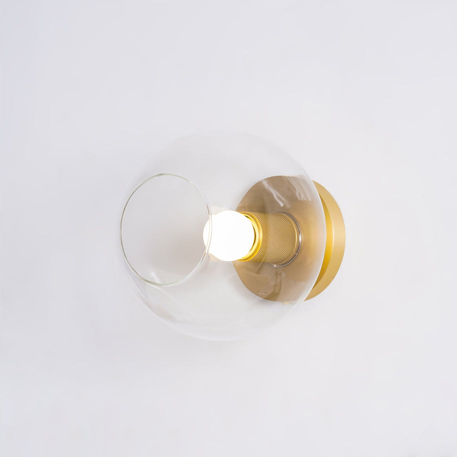 Light fitting with a round brass-toned back plate and a rounded glass shade and an exposed bulb, lit, shown at a slight angle on a pale background.