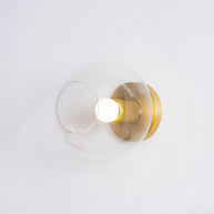 Light fitting with a round brass-toned back plate and a rounded glass shade and an exposed bulb, lit, shown at a slight angle on a pale background.