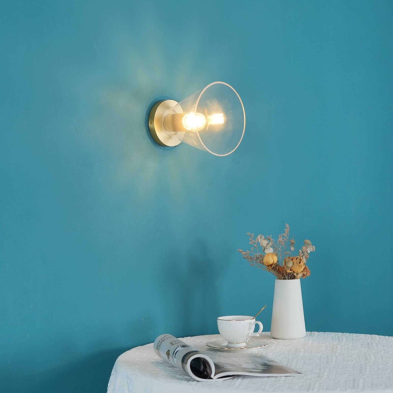 Light fitting with a round brass-toned back plate and a rounded glass shade, lit, against a teal background, seen above a laid table.