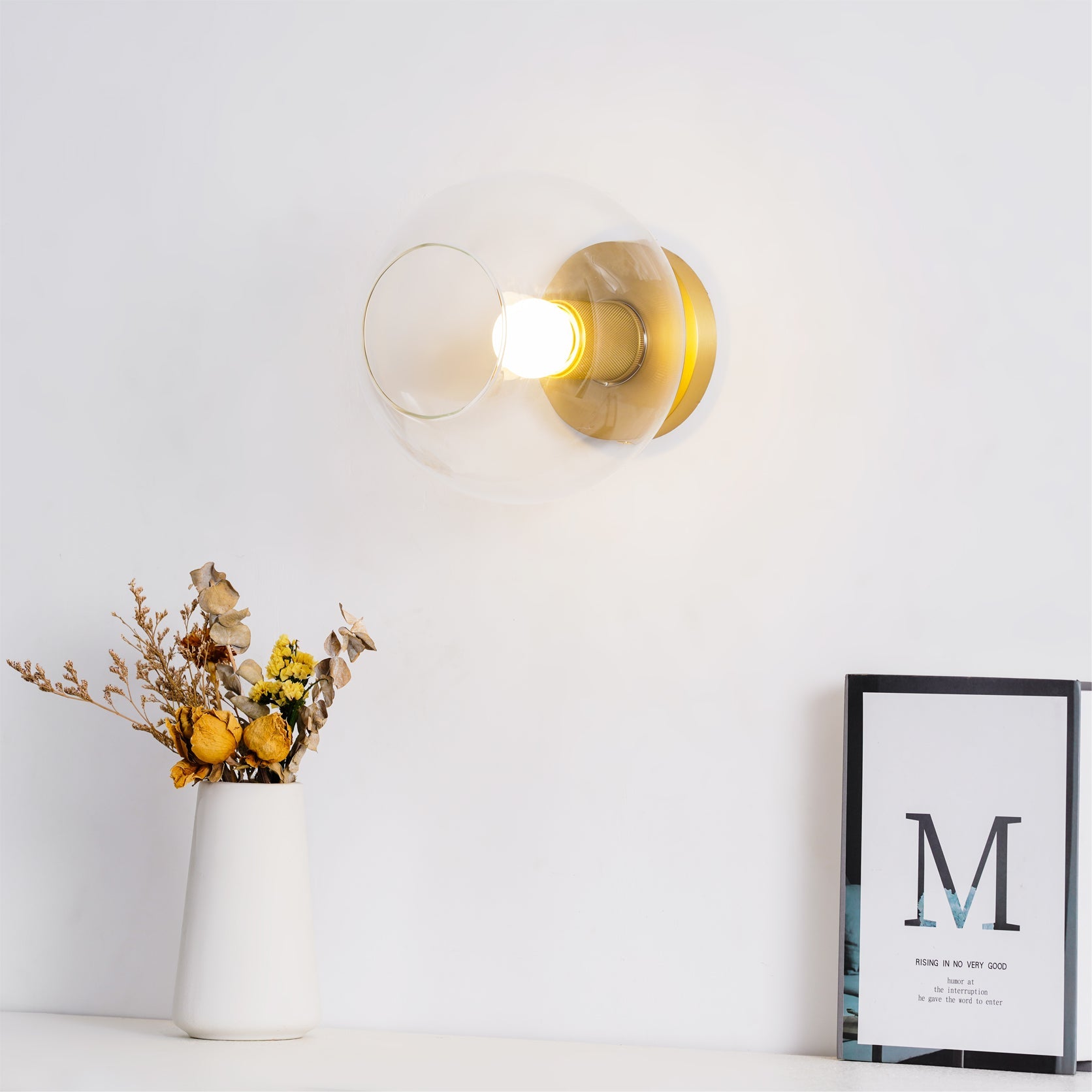 Light fitting with a round brass-toned back plate and a rounded glass shade, lit, against a white background, with dried flowers and a framed card below.