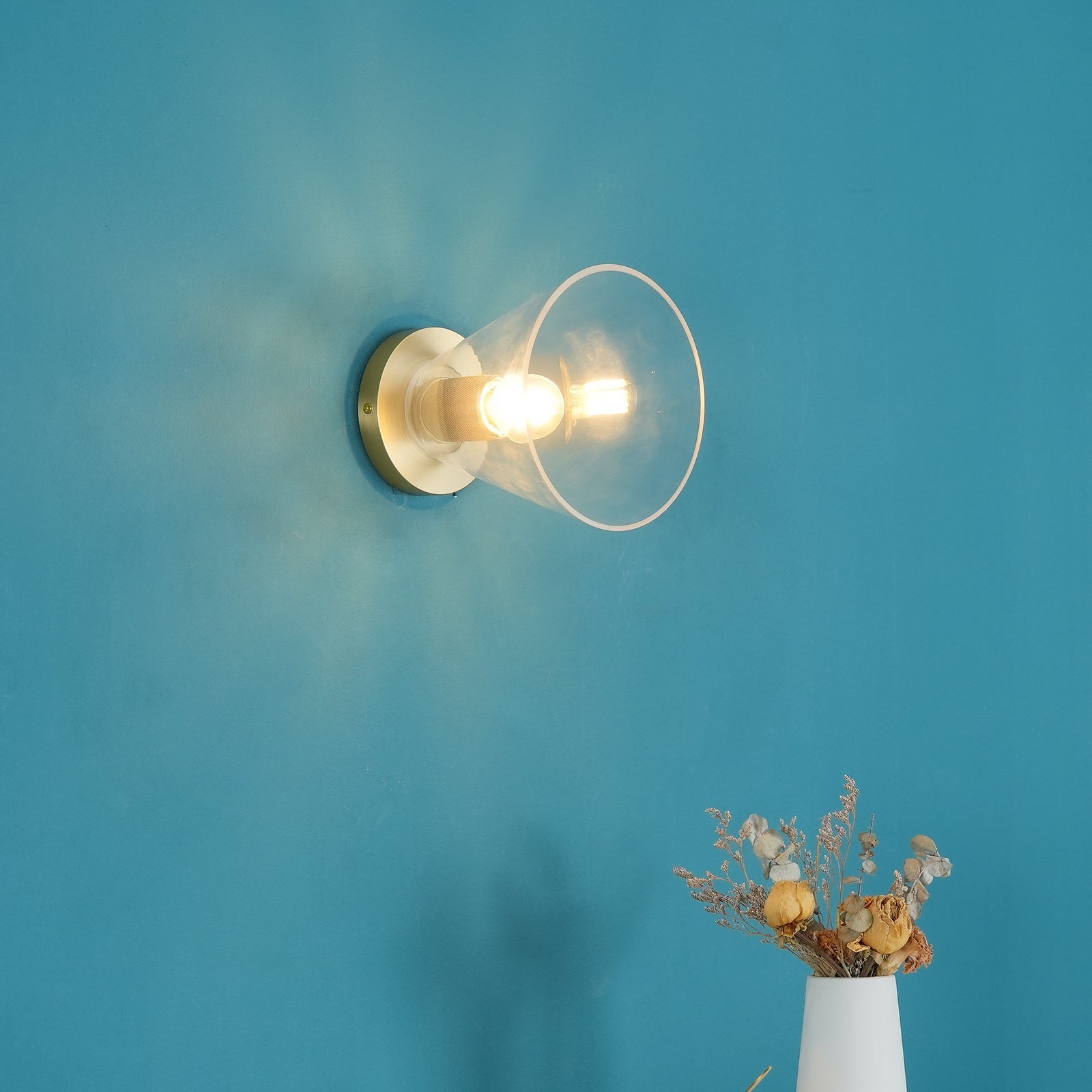 Light fitting with a round brass-toned back plate and a rounded glass shade, lit, against a teal background, seen from further back.