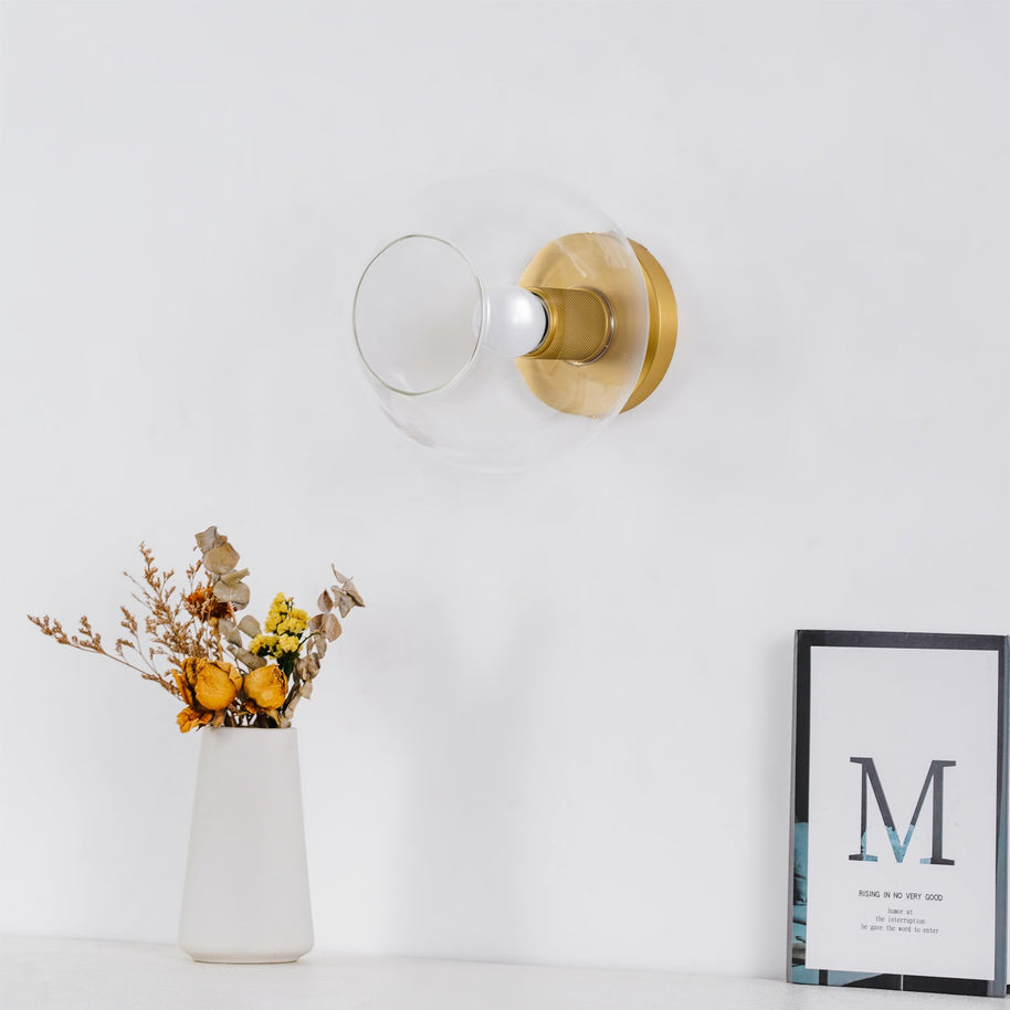 Light fitting with a round brass-toned back plate and a rounded glass shade, lit, against a white background, with a vase of dried flowers and a framed card below.