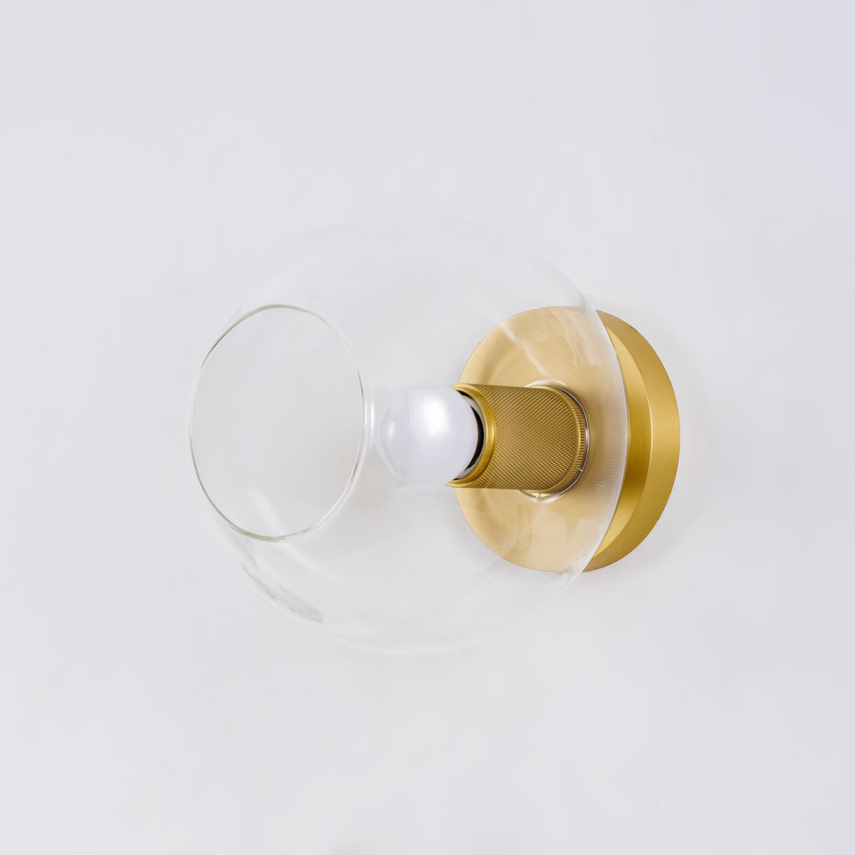 Light fitting with a round brass-toned back plate and a rounded glass shade, lit, against a white background, seen from one side.