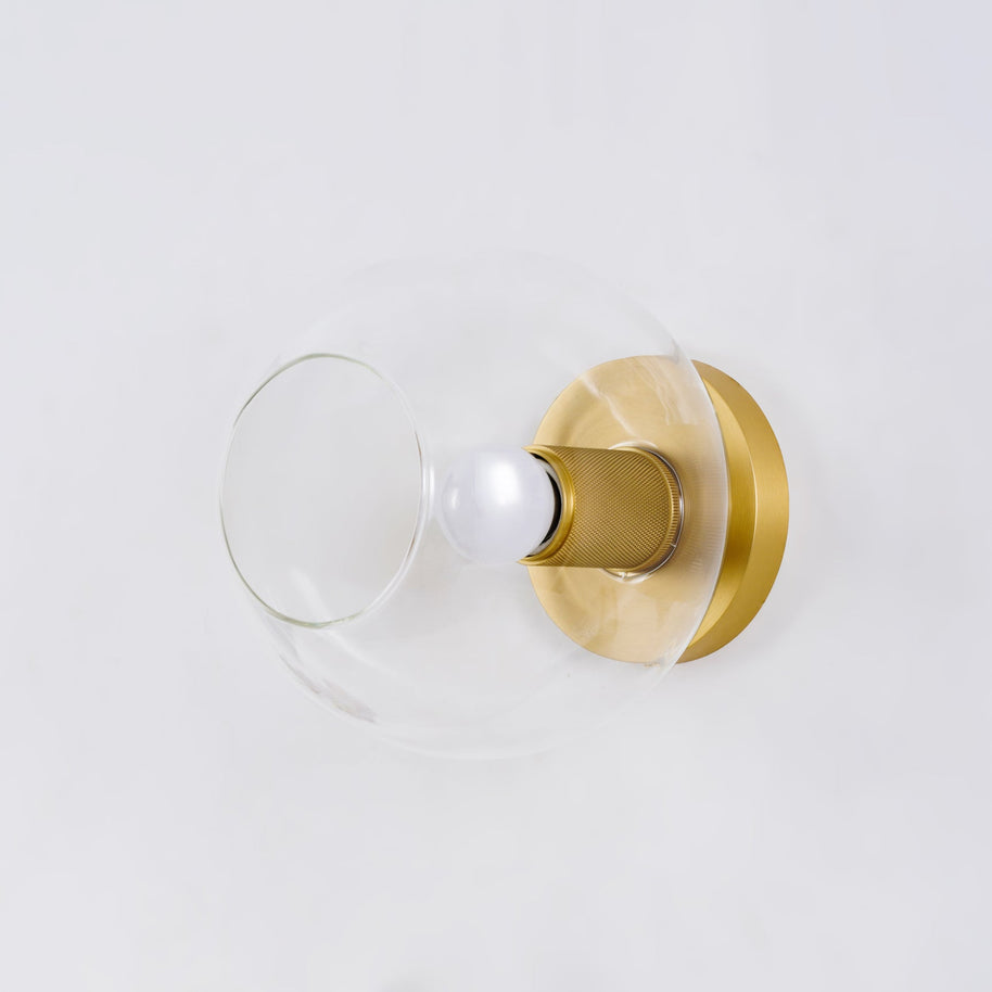 Light fitting with a round brass-toned back plate and a rounded glass shade, lit, against a white background, seen from one side.