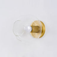Light fitting with a round brass-toned back plate and a rounded glass shade, lit, against a white background, seen from one side.