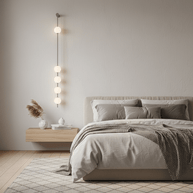 Wall lamp with a vertical row of white ball shades on a slim brass-coloured rod lit beside a bed on a plain wall.