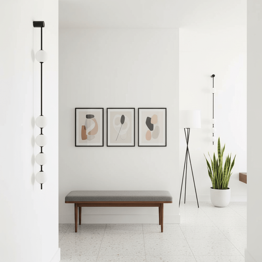 Wall lamp with a vertical row of white ball shades on a slim black rod lit on a white wall beside framed prints.