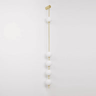 Wall lamp with a vertical row of white ball shades on a slim gold-toned rod, unlit, on a pale wall.