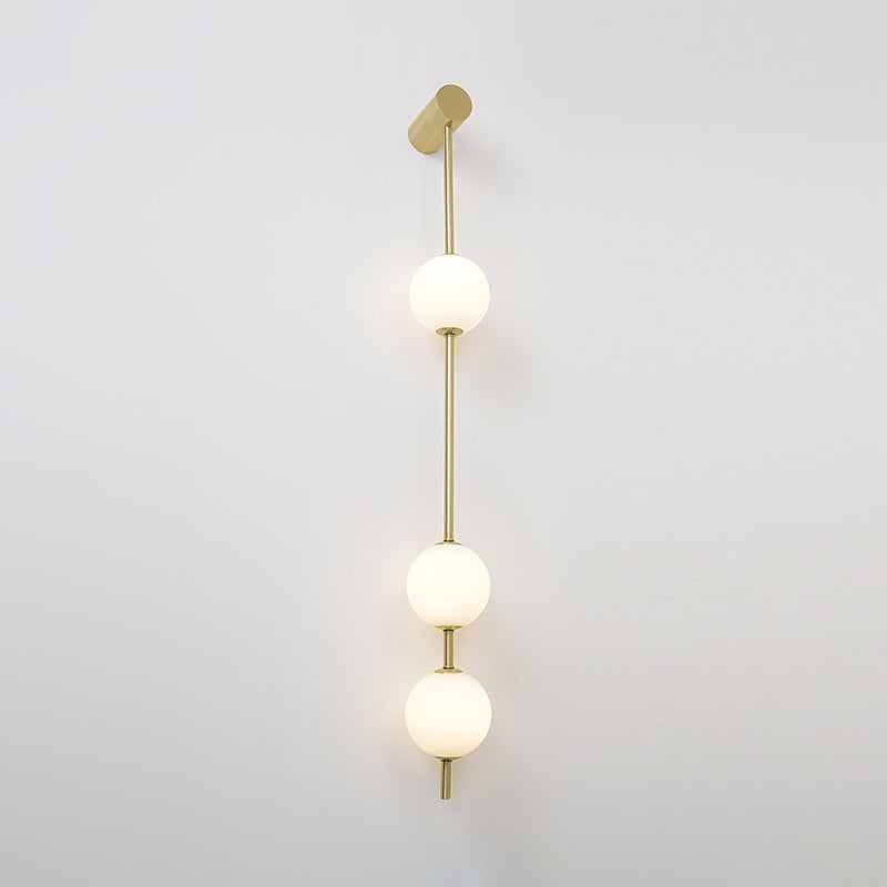 Wall lamp with a vertical row of white ball shades on a slim gold-toned rod, lit, on a pale wall.