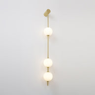 Wall lamp with a vertical row of white ball shades on a slim gold-toned rod, lit, on a pale wall.