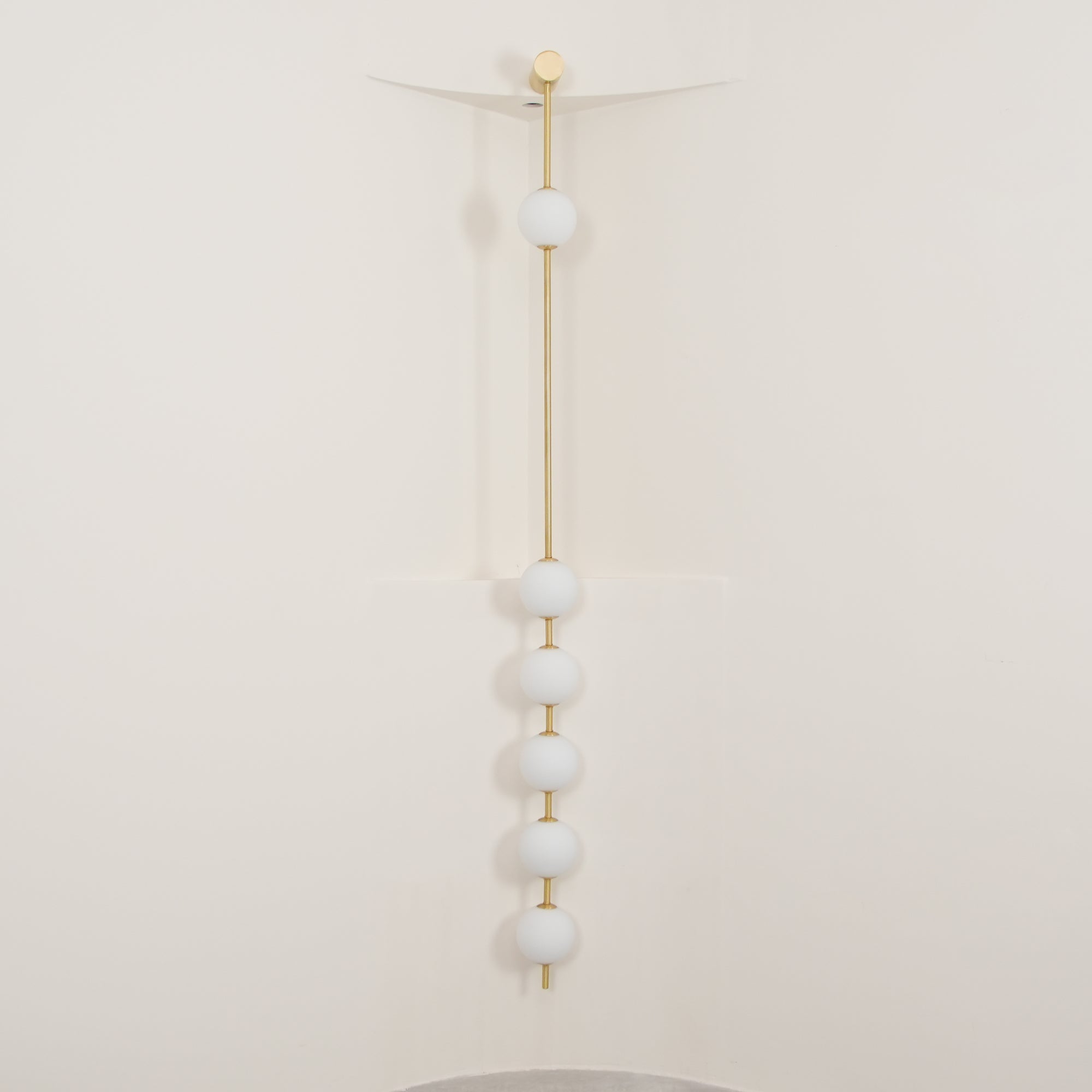 Wall lamp with a vertical row of white ball shades on a slim brass-coloured rod, unlit, against a cream wall.