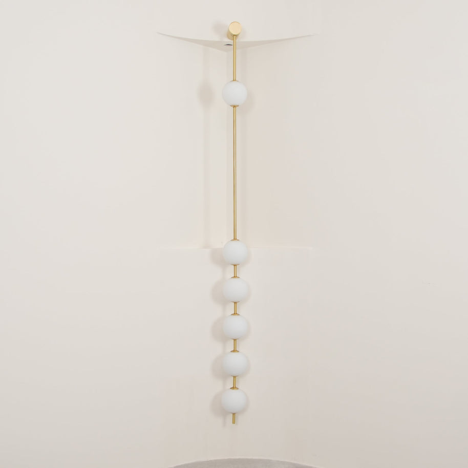 Wall lamp with a vertical row of white ball shades on a slim brass-coloured rod, unlit, against a cream wall.
