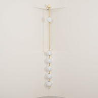 Wall lamp with a vertical row of white ball shades on a slim brass-coloured rod, unlit, against a cream wall.