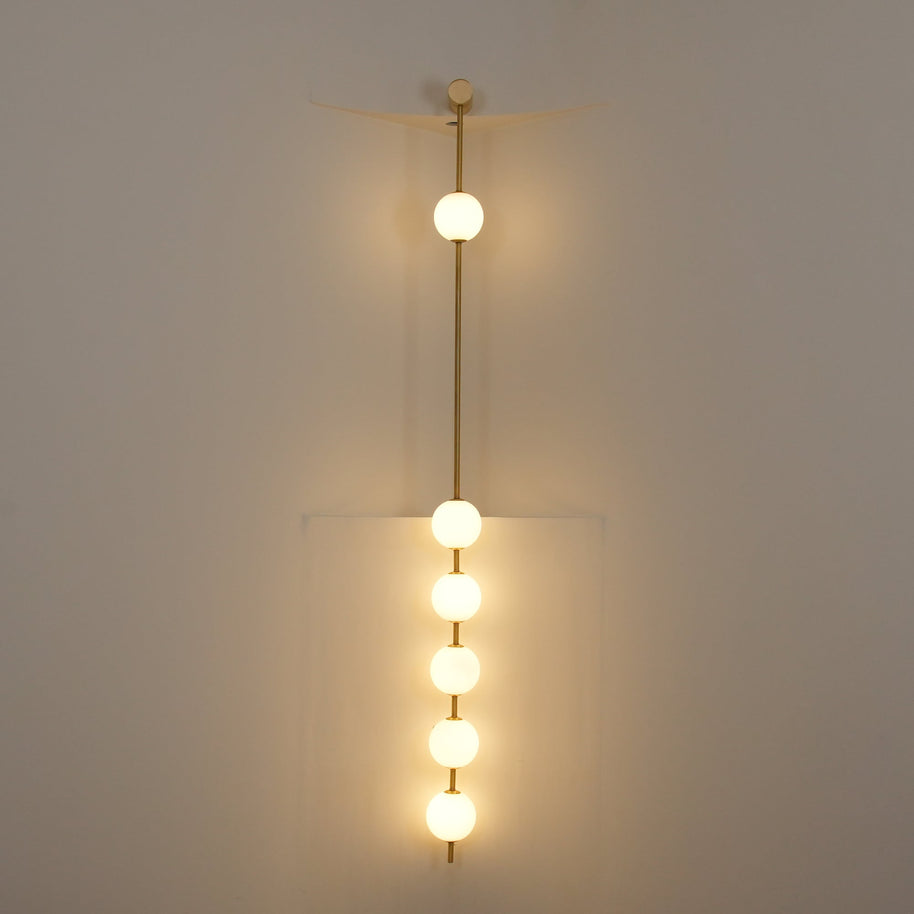 Wall lamp with a vertical row of white ball shades on a slim brass-coloured rod lit against a pale wall, seen close.