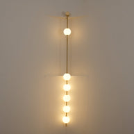 Wall lamp with a vertical row of white ball shades on a slim brass-coloured rod lit against a pale wall, seen close.