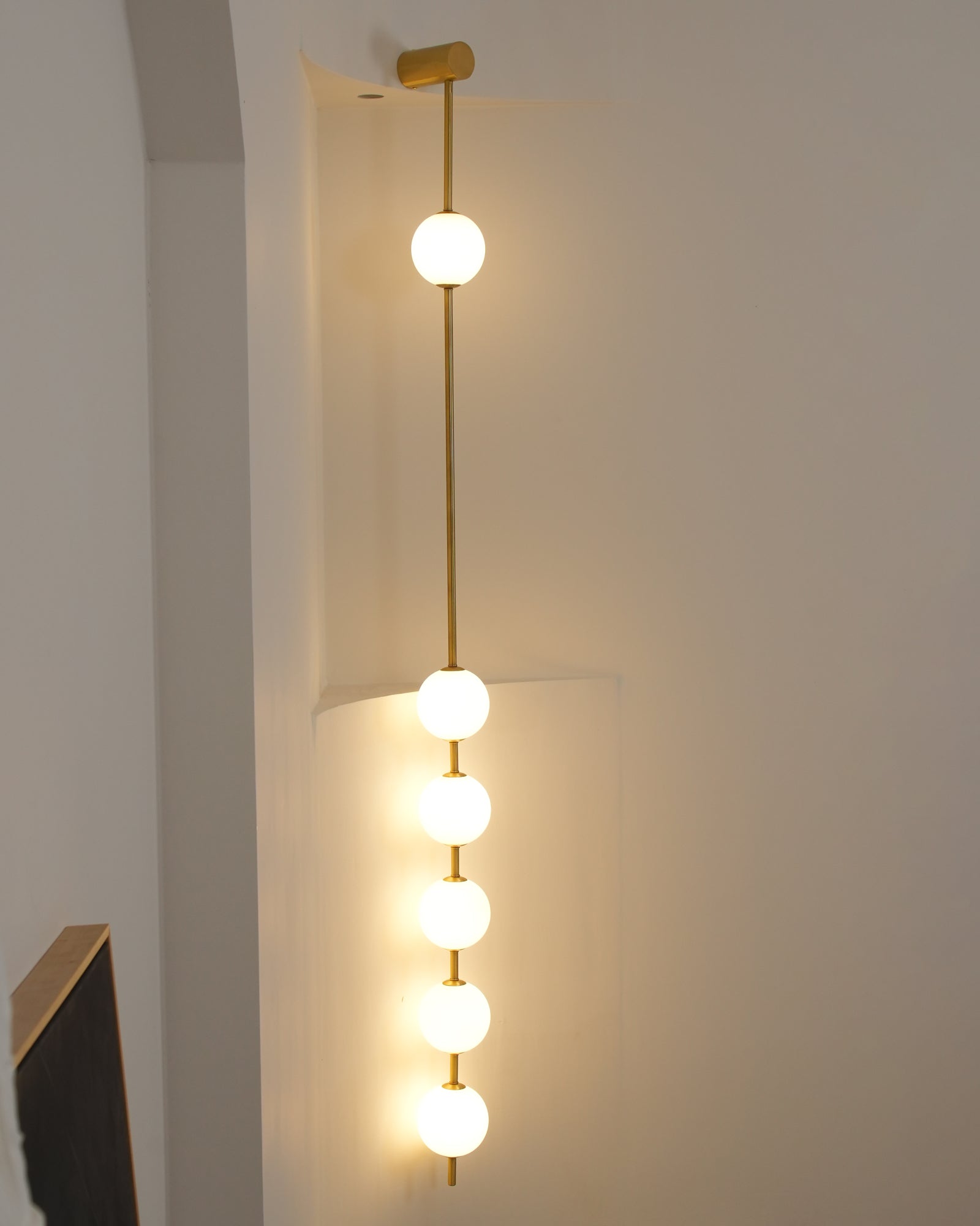 Wall lamp with a vertical row of white ball shades on a slim brass-coloured rod lit against a pale wall.