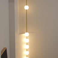 Wall lamp with a vertical row of white ball shades on a slim brass-coloured rod lit against a pale wall.