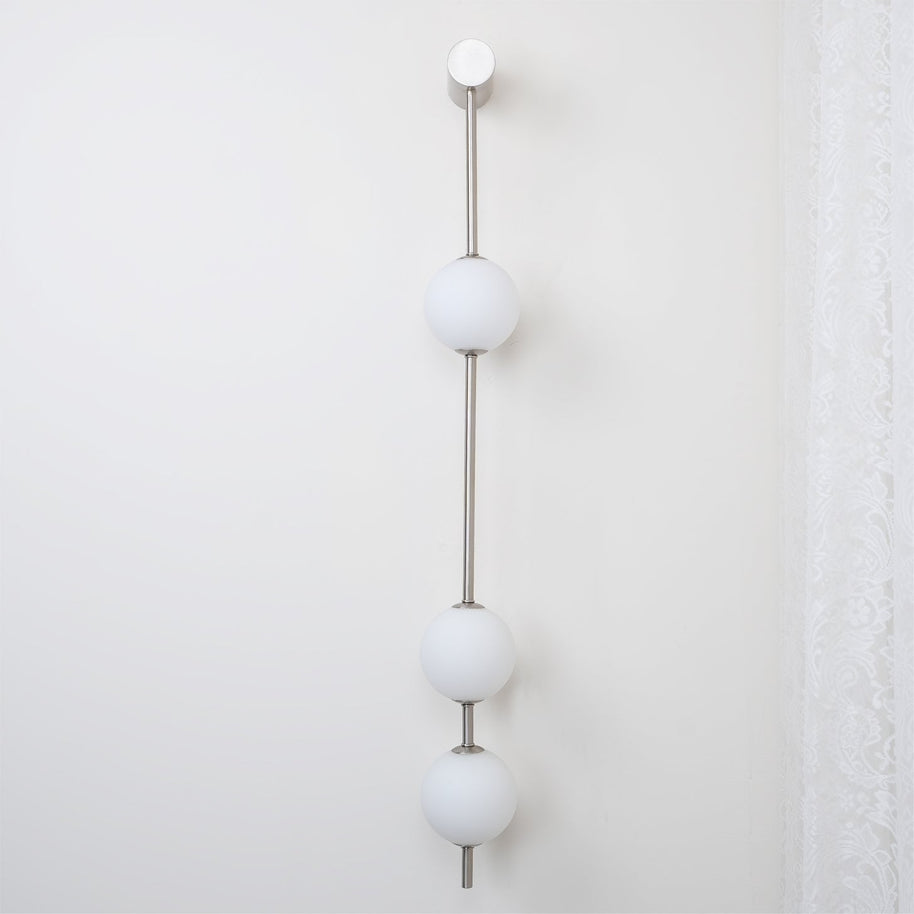 Wall lamp with a vertical row of white ball shades on a slim nickel-coloured rod, unlit, on a white wall beside a curtain.