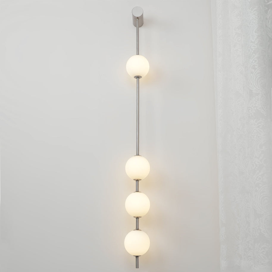 Wall lamp with a vertical row of white ball shades on a slim nickel-coloured rod lit on a white wall beside a curtain.