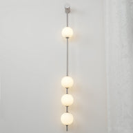 Wall lamp with a vertical row of white ball shades on a slim nickel-coloured rod lit on a white wall beside a curtain.