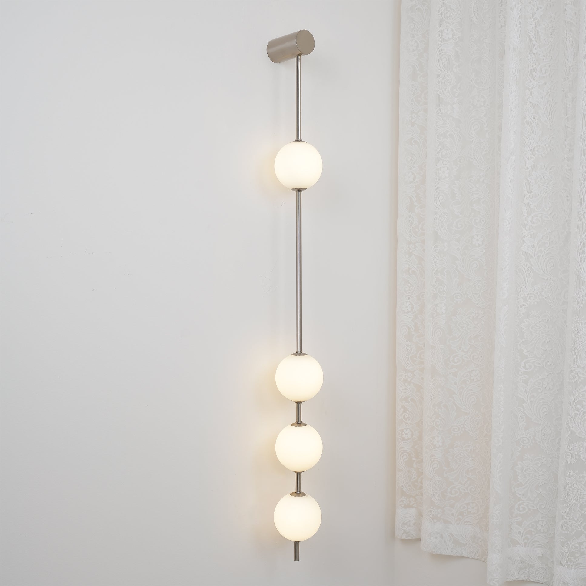 Wall lamp with a vertical row of white ball shades on a slim nickel-coloured rod lit beside a curtain.
