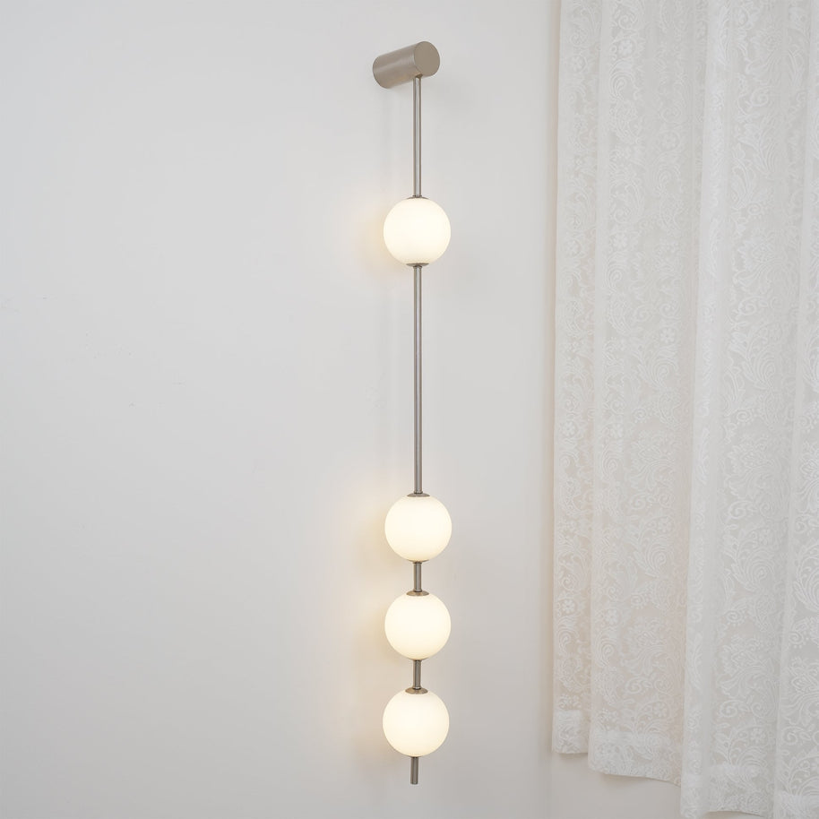 Wall lamp with a vertical row of white ball shades on a slim nickel-coloured rod lit beside a curtain.