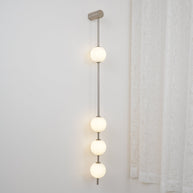 Wall lamp with a vertical row of white ball shades on a slim nickel-coloured rod lit beside a curtain.