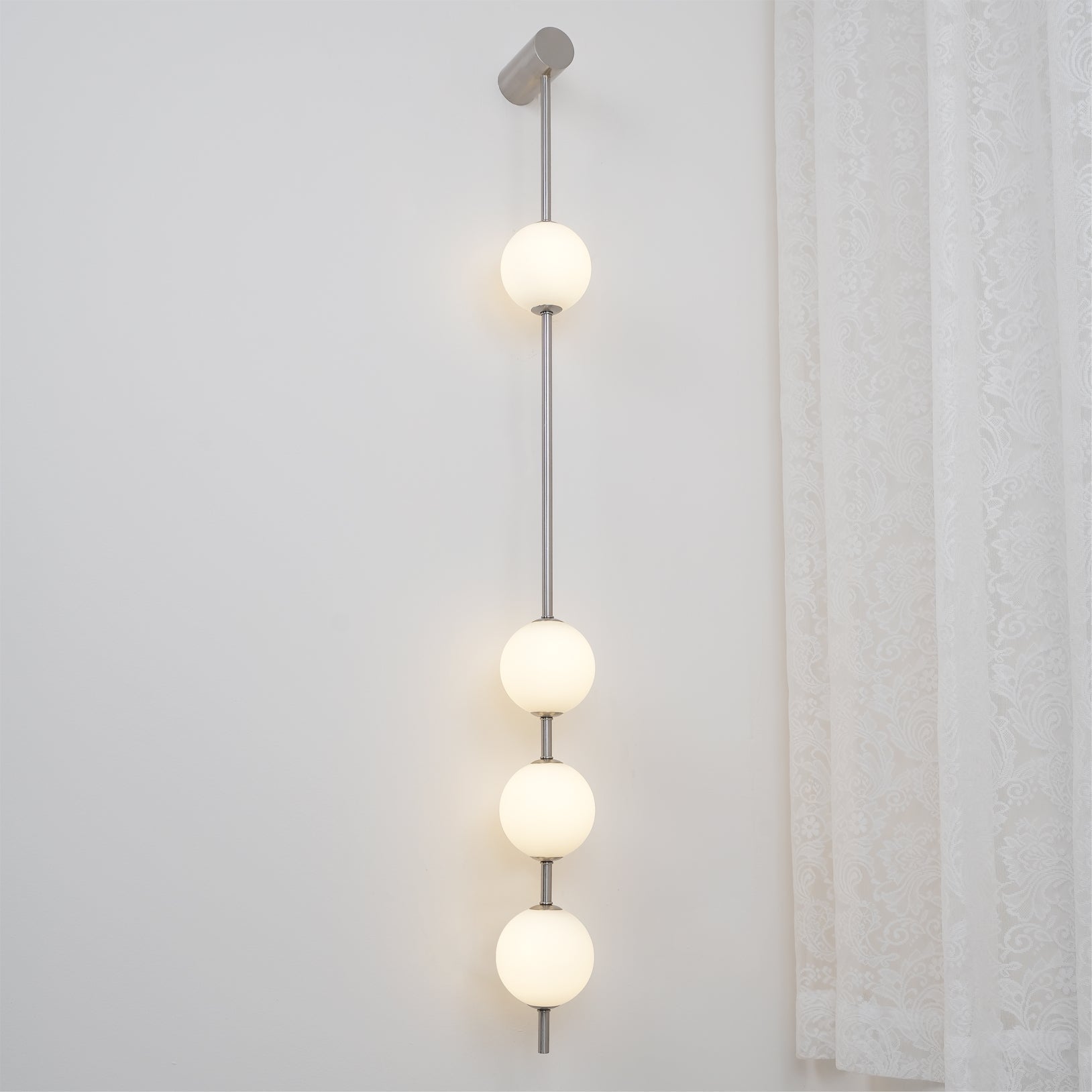 Wall lamp with a vertical row of white ball shades on a slim nickel-coloured rod lit beside a lace curtain.