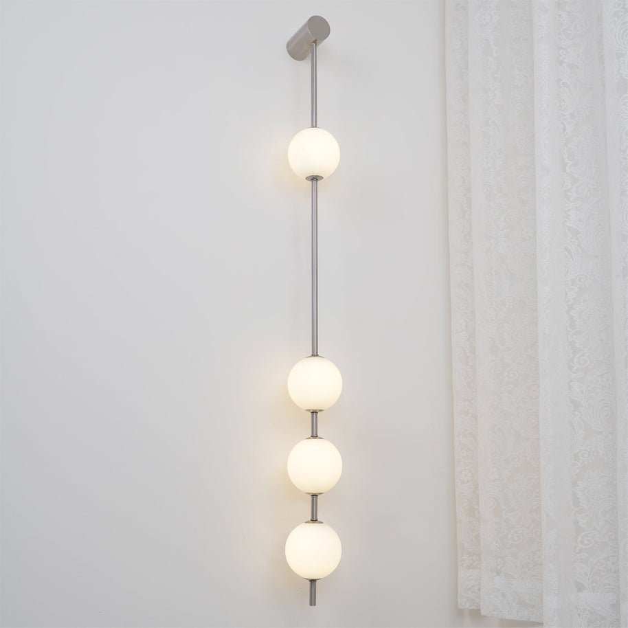 Wall lamp with a vertical row of white ball shades on a slim nickel-coloured rod lit beside a lace curtain.