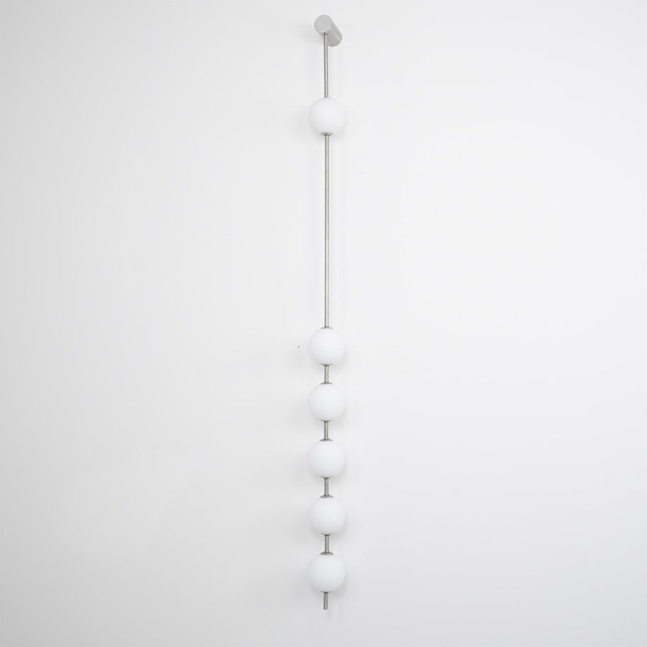Wall lamp with a vertical row of white ball shades on a slim nickel-coloured rod, unlit, on a white wall.