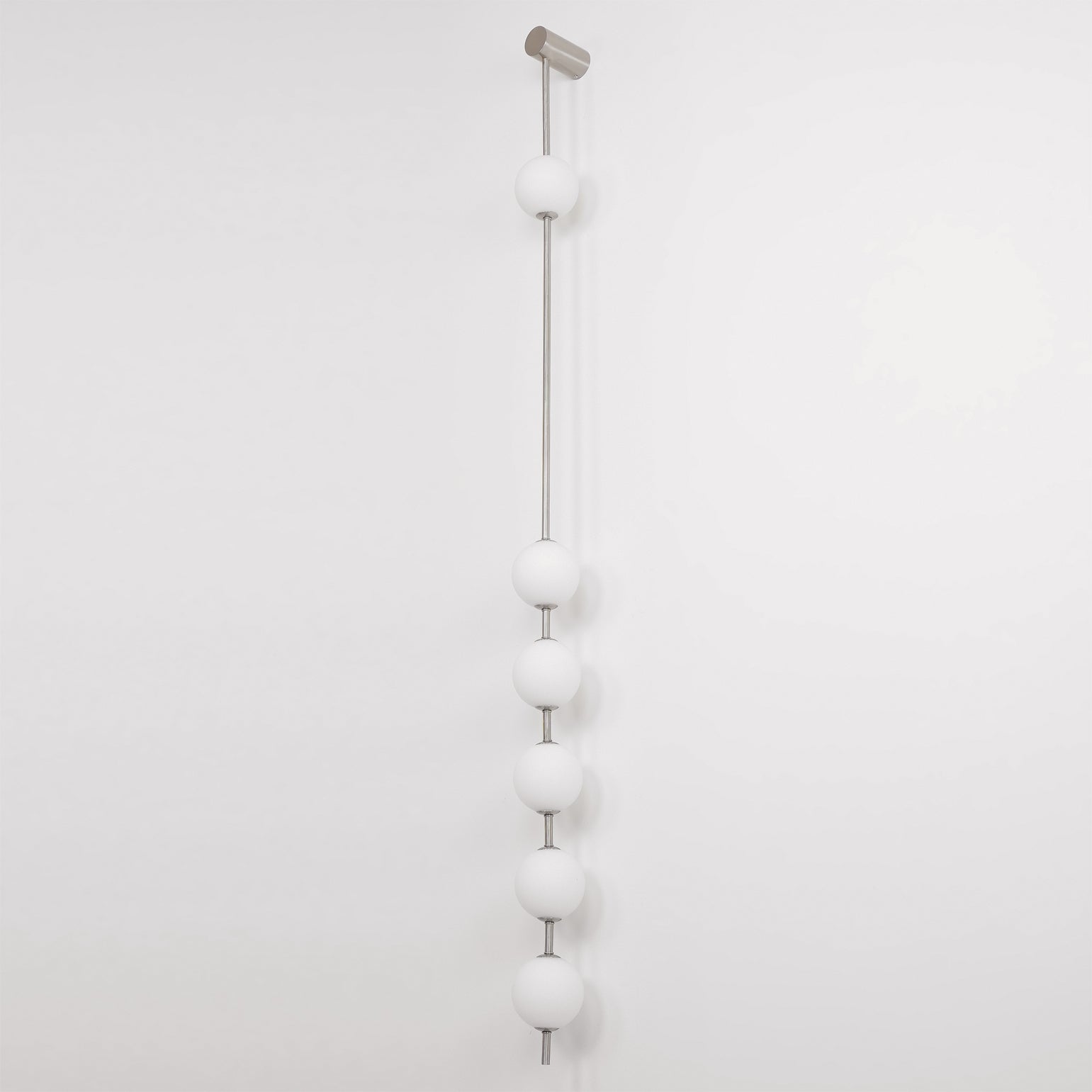 Wall lamp with a vertical row of white ball shades on a slim nickel-coloured rod, unlit, on a white wall.
