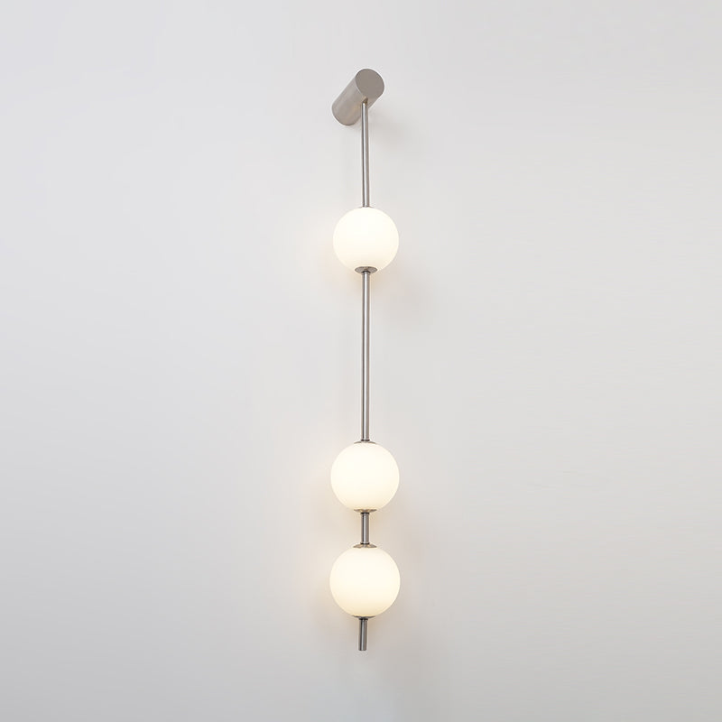 Wall lamp with a vertical row of white ball shades on a slim pale grey rod, lit, on a white wall.