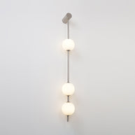 Wall lamp with a vertical row of white ball shades on a slim pale grey rod, lit, on a white wall.