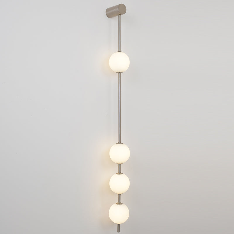 Wall lamp with a vertical row of white ball shades on a slim grey-toned rod, lit, on a grey wall.