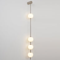 Wall lamp with a vertical row of white ball shades on a slim grey-toned rod, lit, on a grey wall.