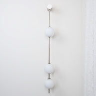 Wall lamp with a vertical row of white ball shades on a slim nickel-coloured rod, unlit, on a white wall beside a curtain.