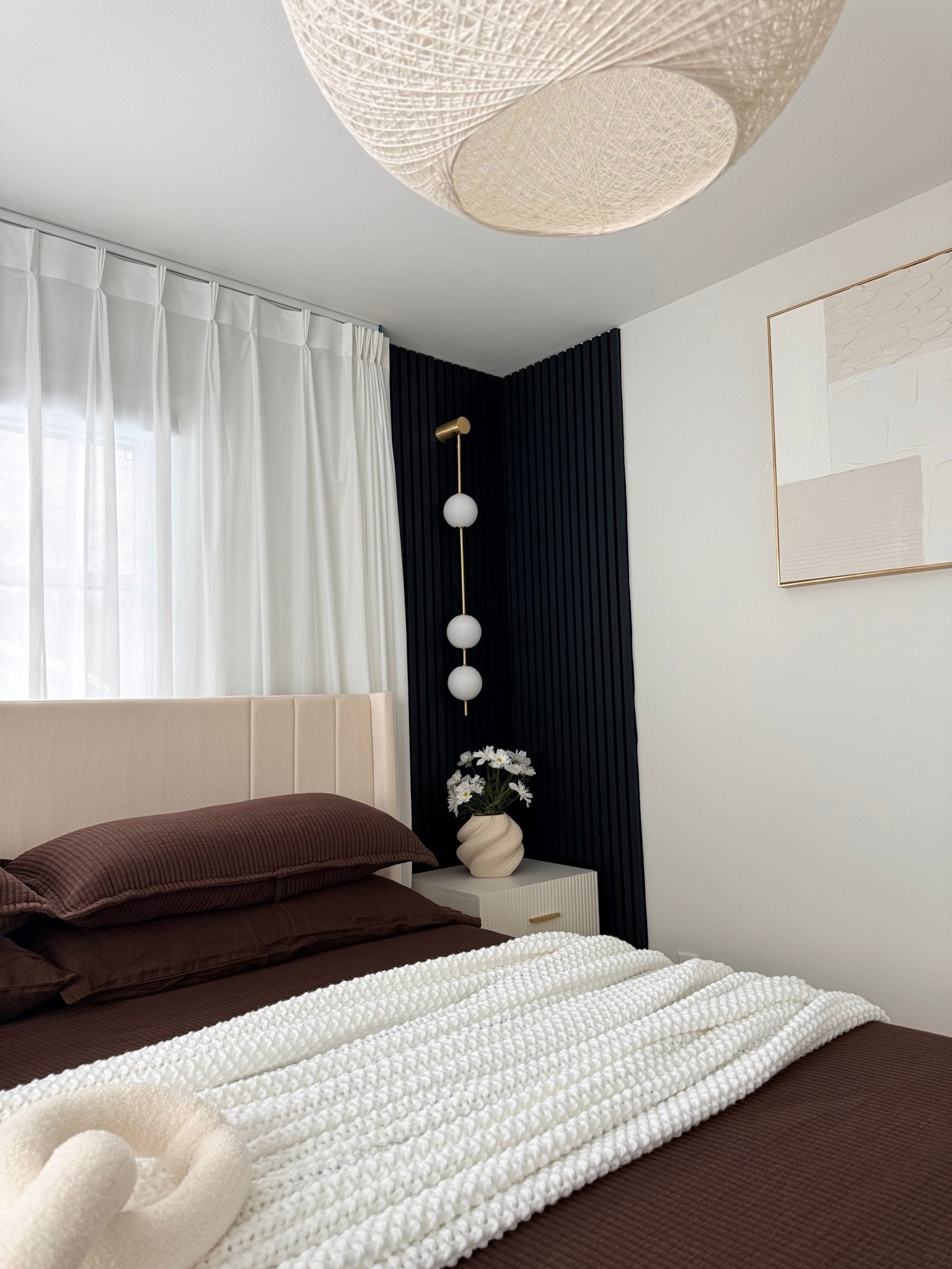 Wall lamp with a vertical row of white ball shades on a slim brass-coloured rod lit on a dark blue panelled wall beside a bed.