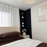 Wall lamp with a vertical row of white ball shades on a slim brass-coloured rod lit on a dark blue panelled wall beside a bed.