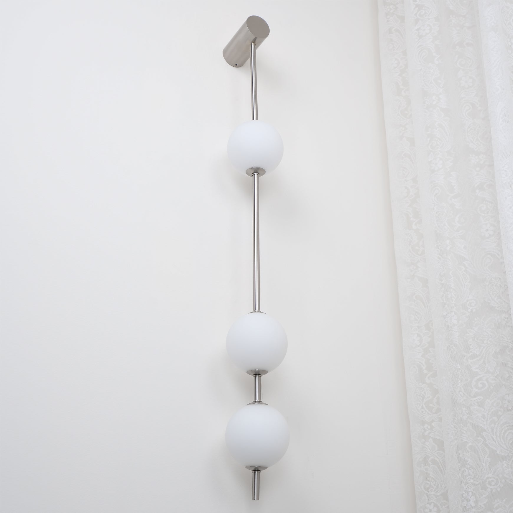 Wall lamp with a vertical row of white ball shades on a slim nickel-coloured rod, unlit, seen close on a white wall.