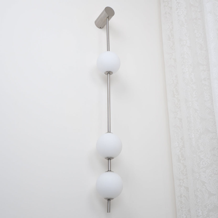 Wall lamp with a vertical row of white ball shades on a slim nickel-coloured rod, unlit, seen close on a white wall.