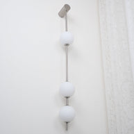 Wall lamp with a vertical row of white ball shades on a slim nickel-coloured rod, unlit, seen close on a white wall.