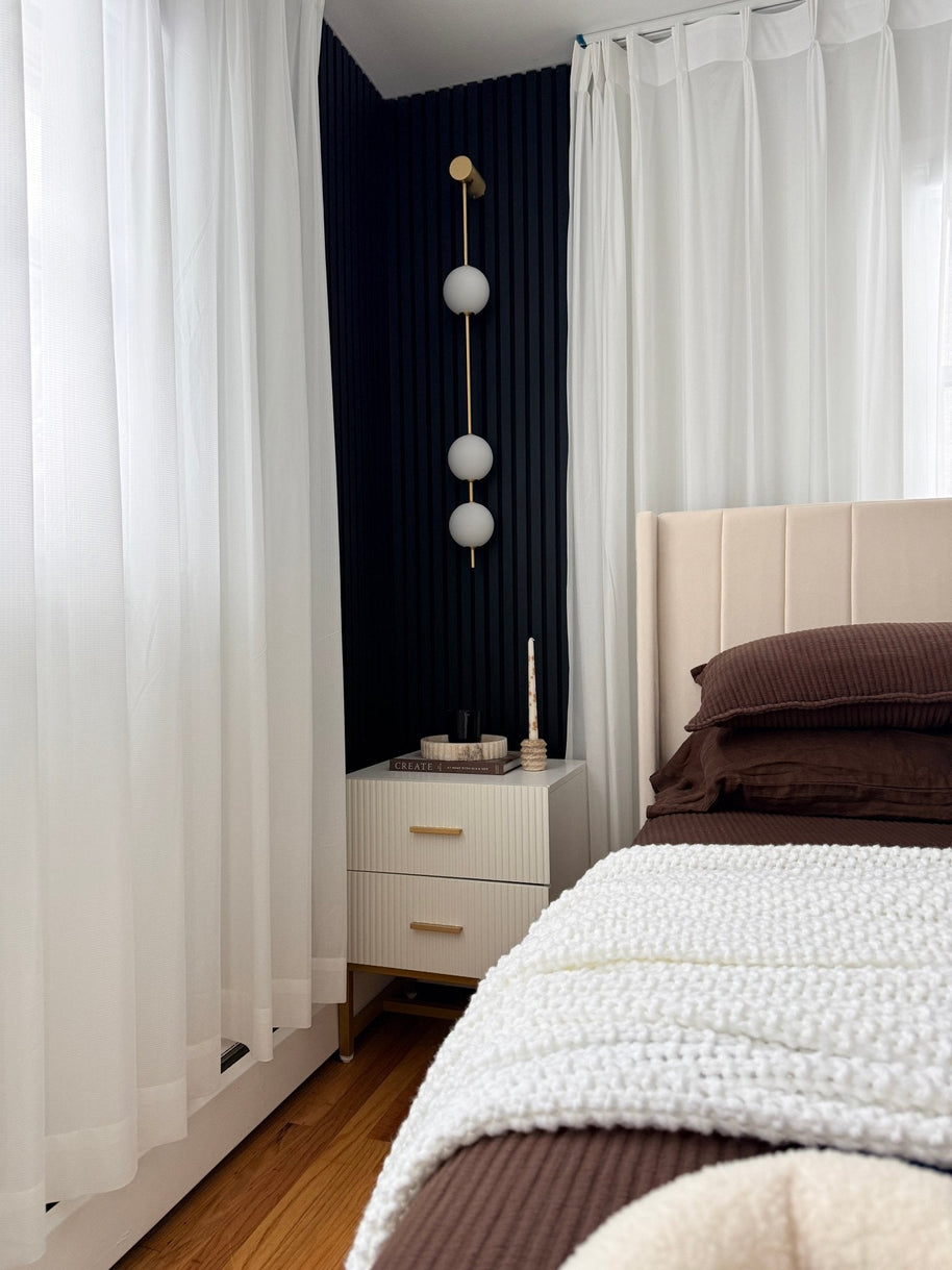 Wall lamp with a vertical row of white ball shades on a slim brass-coloured rod lit beside a bed and a sheer curtain.