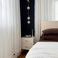 Wall lamp with a vertical row of white ball shades on a slim brass-coloured rod lit beside a bed and a sheer curtain.