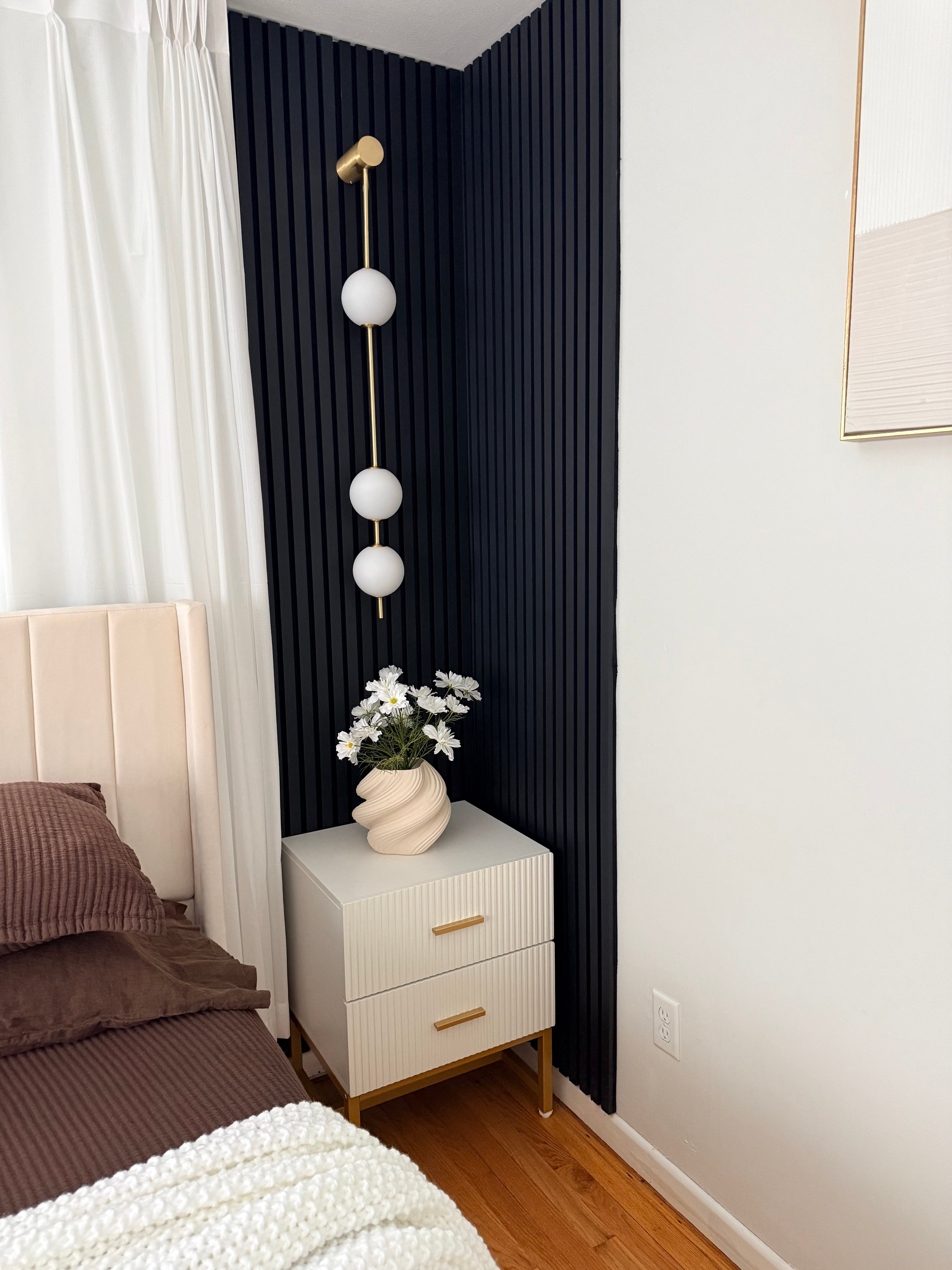 Wall lamp with a vertical row of white ball shades on a slim brass-coloured rod lit on a navy panelled wall beside a bed.