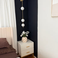 Wall lamp with a vertical row of white ball shades on a slim brass-coloured rod lit on a navy panelled wall beside a bed.