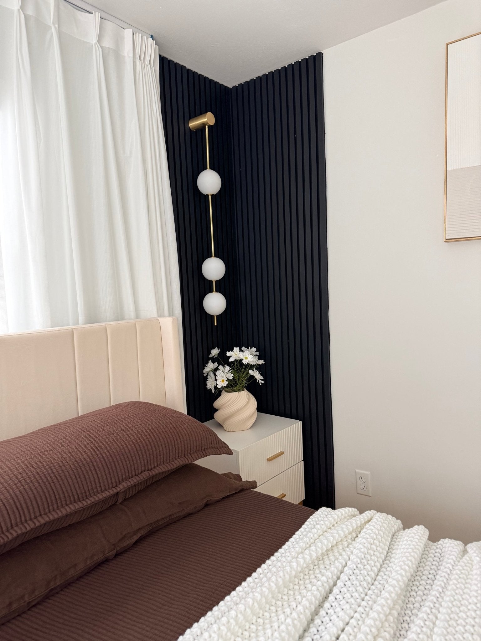 Wall lamp with a vertical row of white ball shades on a slim brass-coloured rod lit in a bedroom corner with navy panelling.