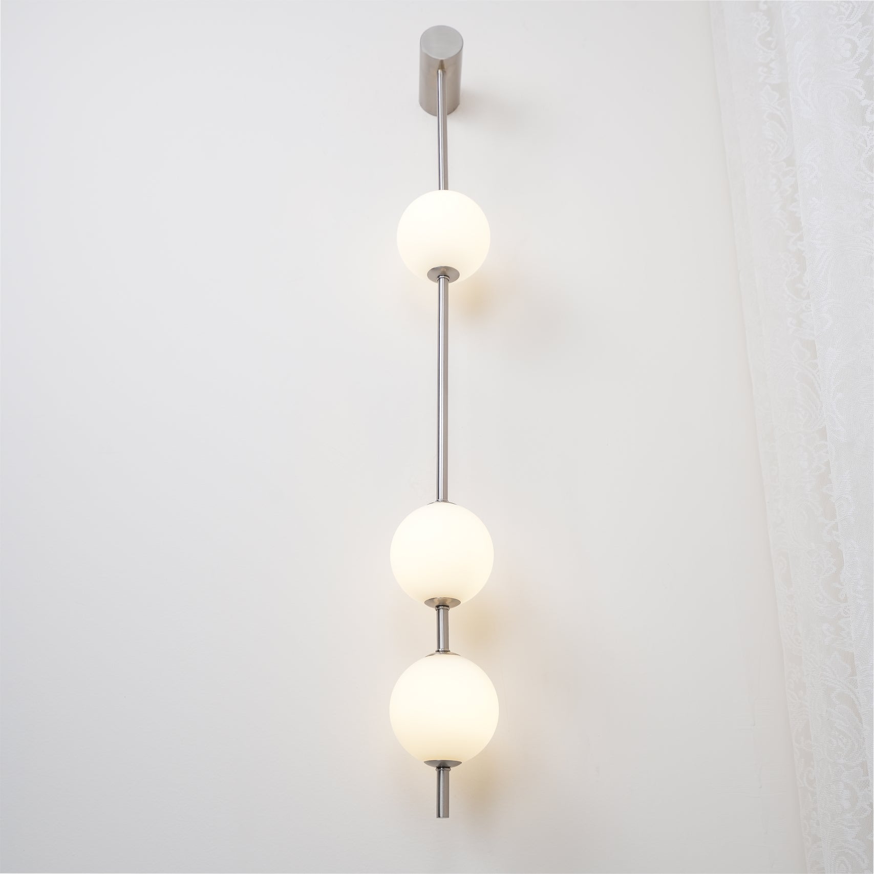 Wall lamp with a vertical row of white ball shades on a slim nickel-coloured rod lit beside a lace curtain.